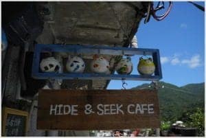 Colorful decorative ceramic figures hanging above a wooden sign reading "HIDE & SEEK CAFE," set against a blue sky and green mountains, emphasizing travel-related dining experiences.