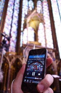 Hand holding smartphone displaying music player interface, set against a backdrop of colorful stained glass windows in a historic church, symbolizing connectivity and cultural exploration while traveling.
