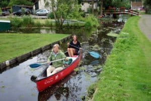Two women paddling in a red canoe on a serene canal, surrounded by lush greenery and quaint houses, highlighting a canoeing adventure.