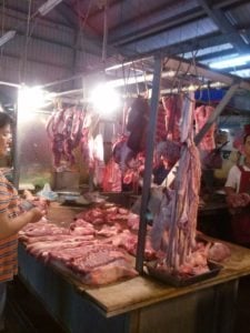 Market stall displaying various cuts of meat with hanging carcasses and a vendor preparing products, highlighting culinary experiences in travel.