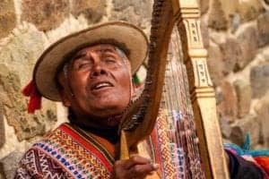 Musician playing a traditional Dutar, wearing colorful attire and a hat, against a stone wall backdrop, representing cultural heritage in travel experiences.