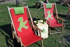Two red deck chairs with green accents featuring a bear logo, placed on grass with a small table holding two bottles, representing a relaxed outdoor setting for travelers.