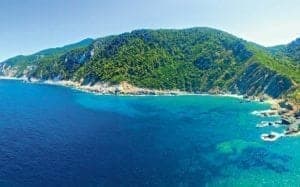 Chapel of St. John the Baptist nestled among lush greenery on a coastal hillside, with clear blue waters in the foreground.