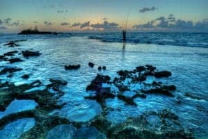 Scenic view of rocky shoreline at sunset in Hawaii, featuring calm waters and a lone fisherman against a colorful sky, emphasizing travel inspiration for Hawaii.