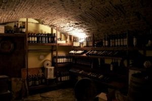 Wine cellar with wooden shelves filled with bottles, dimly lit by overhead light, showcasing a rustic ambiance ideal for exploring wine tasting experiences.