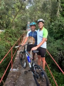 Two cyclists posing on a narrow bridge surrounded by lush greenery, showcasing cross-country cycling adventure.
