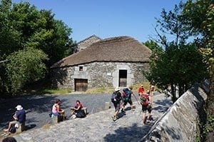 Pilgrims resting near a traditional stone building along the Camino de Santiago, surrounded by greenery and a clear blue sky.