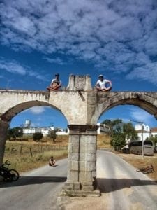 Two people sitting on an old stone bridge with arches, surrounded by rural scenery, illustrating a unique bridge-tunnel experience for travelers.