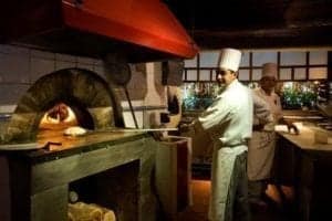 Chefs in a restaurant kitchen preparing food, with a traditional stone oven and culinary activity, highlighting Gustino's focus on travel and culinary experiences.