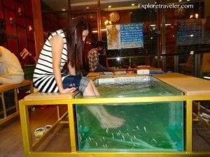 Woman sitting on the edge of a fish spa tank, dipping her feet into the water, with a vibrant indoor setting and other patrons in the background, illustrating unique travel experiences.