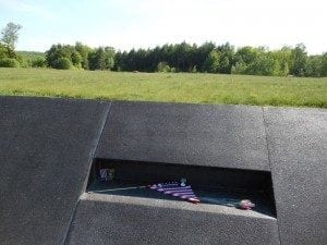 9/11 Memorial site featuring a black granite wall with a small opening displaying a folded American flag and flowers, surrounded by a serene green landscape, symbolizing remembrance and tribute to the victims of the September 11 attacks.