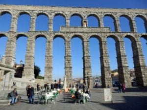 Aqueduct of Segovia with towering stone arches, bustling plaza with visitors and outdoor seating, showcasing historical architecture and vibrant atmosphere.