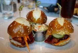 Three gourmet cheeseburgers on a plate, featuring pretzel buns, with a side of coleslaw, showcasing ExploreTraveler's focus on unique food experiences.