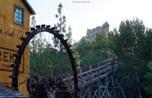 Old wooden water wheel beside a rustic building and a wooden bridge, surrounded by trees and rocky hills, representing travel experiences and outdoor adventures.