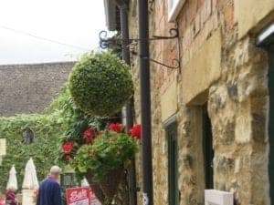 Charming stone facade adorned with greenery and vibrant red flowers, featuring a hanging spherical topiary and a glimpse of outdoor seating, enhancing the travel atmosphere.