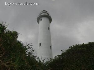 White lighthouse at Da-bai-sha in Taiwan, surrounded by green foliage and under a cloudy sky.