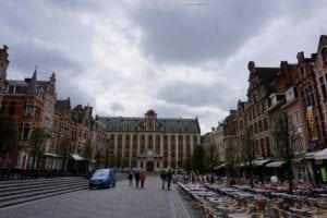 View of the Singel area in Belgium, featuring historic buildings, a central square with people walking, and outdoor seating at cafes under a cloudy sky.