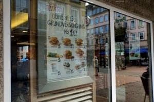 Hamburger promotional poster displayed in a restaurant window, featuring various burger options and prices, highlighting McDonald's offerings in a travel-focused context.