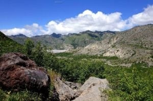 Shrubland landscape near Mount St. Helens featuring rocky terrain, green vegetation, and distant mountains under a blue sky with clouds.