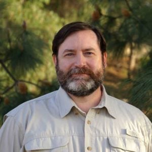 Man with a beard wearing a light-colored shirt, standing outdoors among pine trees, representing travel experiences and cultural insights.
