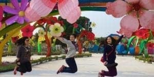 Three women jumping joyfully in a vibrant floral park, surrounded by large decorative flowers and colorful arches, representing a lively travel experience.
