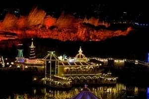 Disneyland Park at night, featuring festive lights and decorations, with illuminated attractions and a colorful backdrop of mountains, showcasing the park's holiday atmosphere.