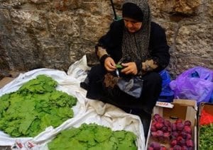 Woman preparing grape leaves at a market, surrounded by fresh greens and produce, highlighting culinary practices related to greens in international cuisines.