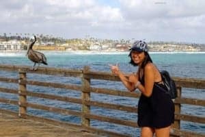 Woman smiling and pointing at a pelican on a wooden pier overlooking the ocean in Oceanside, California, highlighting the beach atmosphere.