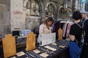 Woman selling handmade jewelry at a market stall with clothing rack in background, showcasing ExploreTraveler experience in Lisbon.
