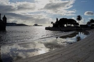 Scenic view of the Belem Tower waterfront in Lisbon, Portugal, featuring ornamental decor, reflecting water, and palm trees under a cloudy sky.