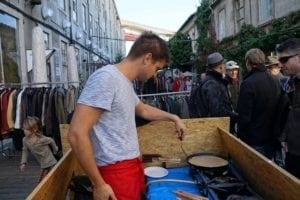 Man preparing street food at a market during SteemFest in Lisbon, surrounded by attendees and clothing stalls, highlighting the vibrant food culture.
