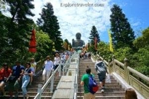 Visitors ascending stairs towards the Big Buddha statue in Hong Kong, surrounded by lush greenery and colorful flags, capturing the essence of tourism and exploration.