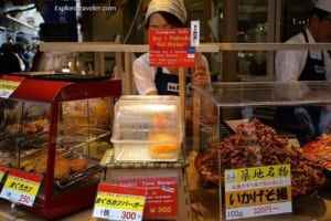 Market stall in Japan showcasing various traditional foods, including fried items and seafood, with signage in Japanese and a vendor in the background, emphasizing Japan's unique culinary culture.