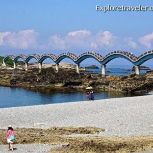 Taiwan Dragon Bridge spanning rocky shoreline with beachgoers, showcasing the iconic architectural design against a clear blue sky.