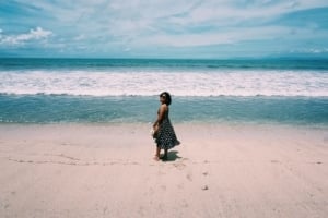 Woman in a black dress standing on a sandy beach, with waves and a blue sky in the background, reflecting ExploreTraveler's travel theme.