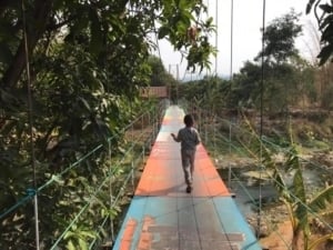 Child walking on a colorful suspension bridge connecting Pasirmuncang and Cikamarang villages in Majalengka, West Java, Indonesia, surrounded by lush greenery.