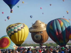 Colorful hot air balloons at the Albuquerque Balloon Fiesta, featuring a bear-shaped balloon with a hat labeled "SMOKEY," amidst a crowd of spectators enjoying the event.
