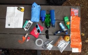 Backpacking essentials for Iceland, including maps, water bottles, snacks, sunscreen, sunglasses, and a multi-tool, arranged on a wooden table.