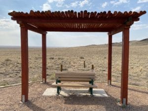 Resting area along the Volcanoes Trail at Petroglyph National Monument, featuring a shaded wooden structure and bench with expansive desert landscape in the background.