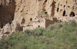 Old pueblo house nestled against rocky cliff at Bandelier National Monument, surrounded by green vegetation.
