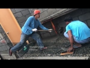 Two men working on construction, one shoveling gravel and the other measuring, with a partially built wall and door in the background, related to Filipino traditional lifestyle and construction methods.