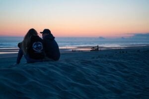 Couple sitting on the sandy beach at Ft. Stevens State Park during an evening sunset, with soft waves and a colorful sky in the background.