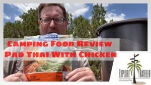 Man holding camping food package labeled "Pad Thai with Chicken," outdoors with greenery in the background, featuring ExploreTraveler logo and text overlay stating "Camping Food Review."