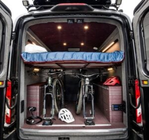 Interior of a camper van featuring a bed above two mountain bikes, with helmets and storage compartments, highlighting travel trends for outdoor adventures in the USA for 2023.