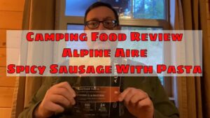 Person holding a camping food package labeled "Alpine Aire Spicy Sausage With Pasta," featuring text overlay "Camping Food Review," set in a cozy indoor environment.