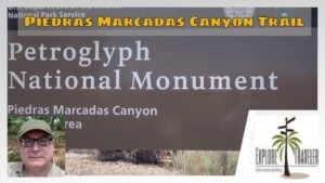 Sign for Petroglyph National Monument at Piedras Marcadas Canyon Trail, featuring park name and trail information, with a person in the foreground.